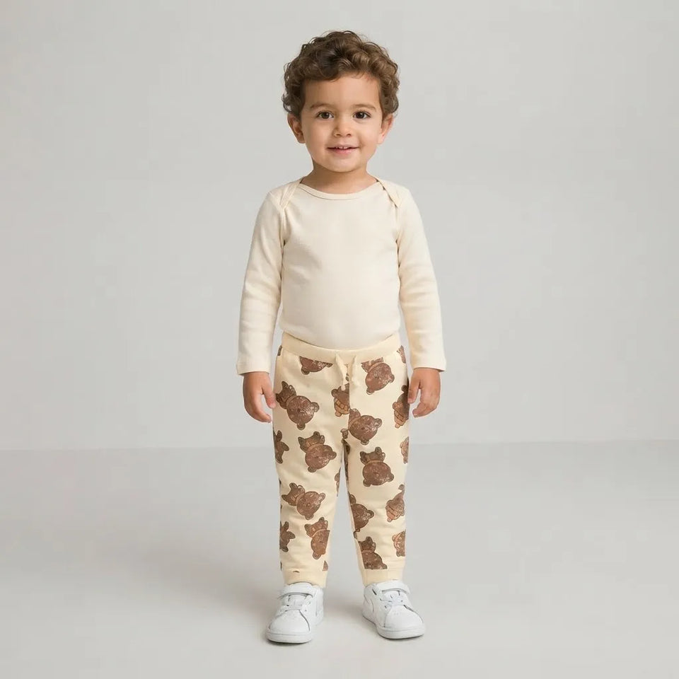 A toddler standing against a light gray studio background, wearing a cream long-sleeve top and matching cream pants with a brown teddy bear print, plus white sneakers.