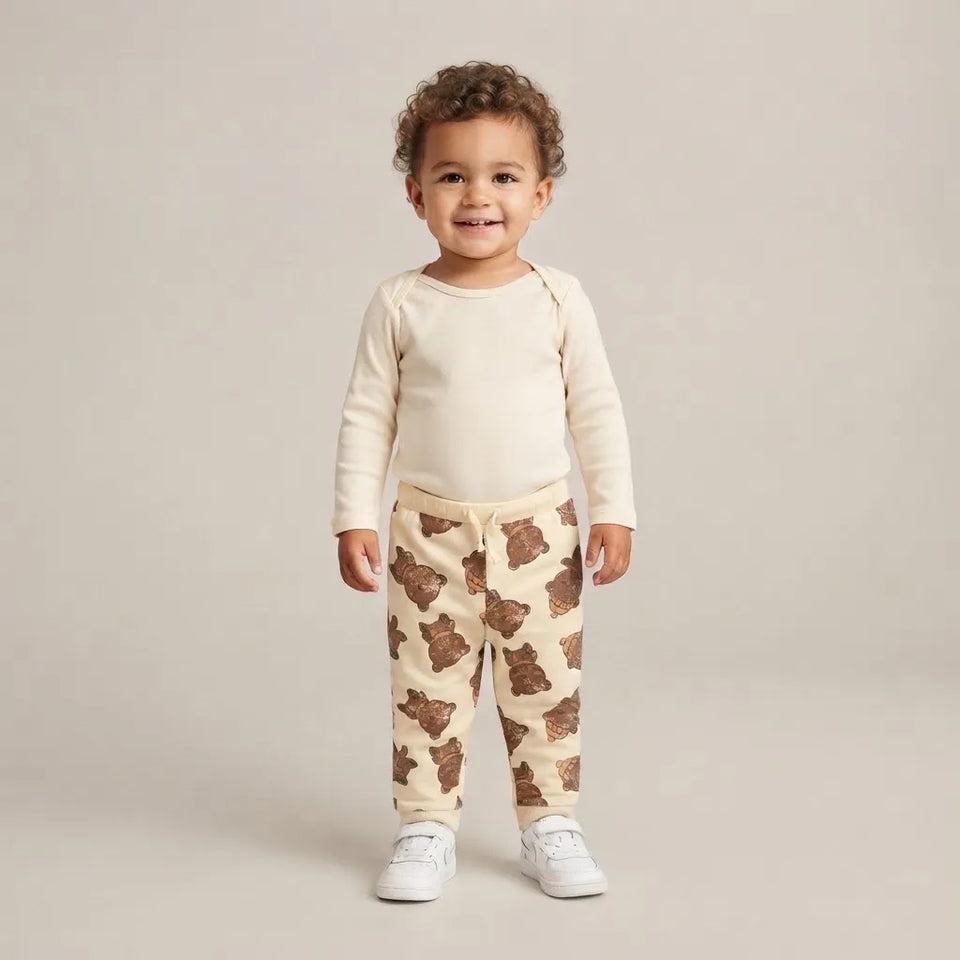 A toddler standing against a light beige studio background, wearing a cream long-sleeve top and matching cream pants with a brown teddy bear print, paired with white sneakers.