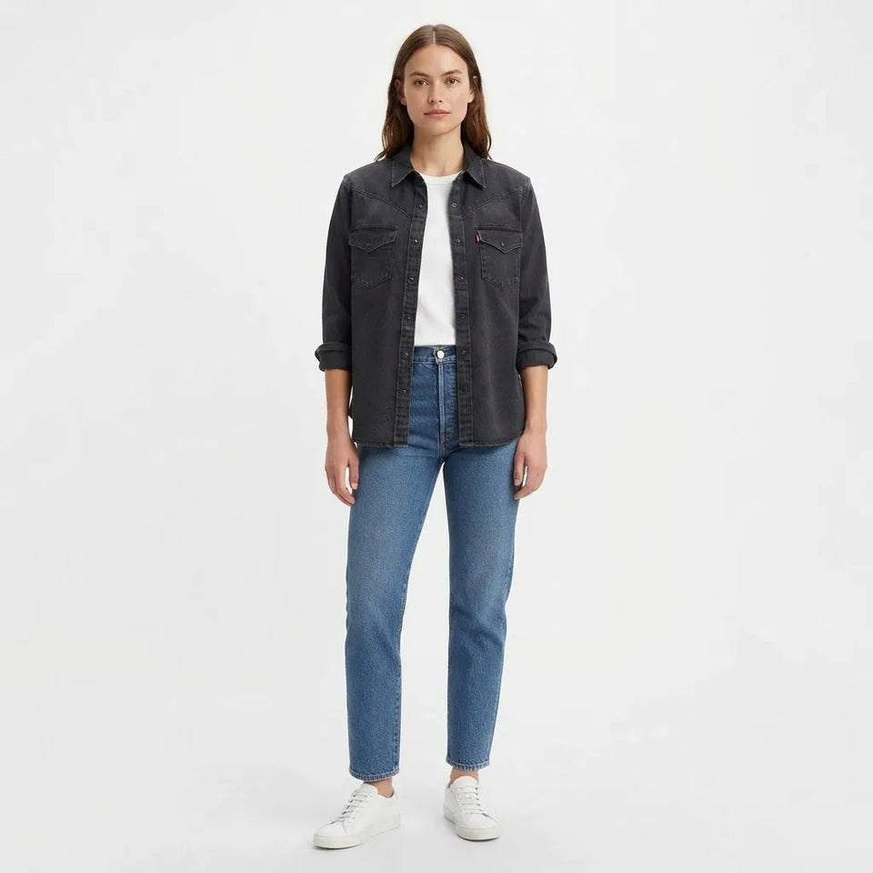 A woman modeling a dark gray denim button-up shirt jacket over a white t-shirt, paired with blue jeans and white sneakers, against a plain white studio background.
