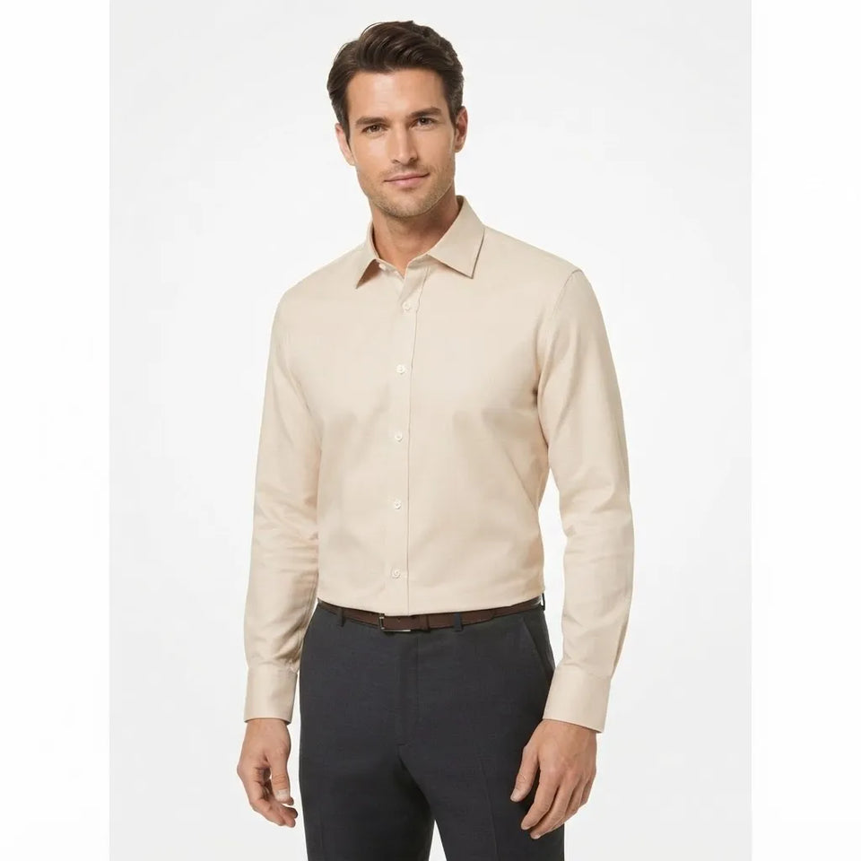 A man wearing a light beige long-sleeve button-down dress shirt with dark gray trousers, standing against a plain white studio background.