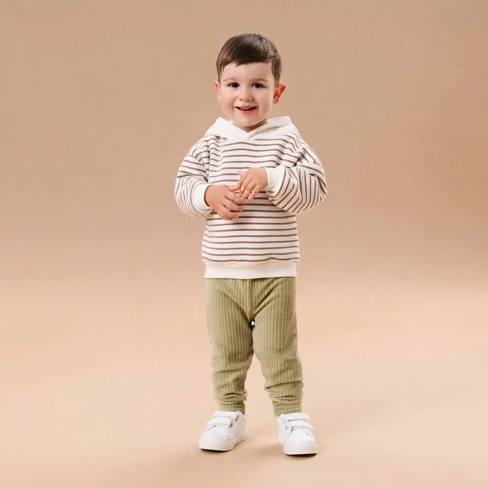 A toddler standing against a solid beige studio background, wearing a cream and brown horizontally striped long-sleeve top with a white collar, olive green ribbed pants, and white sneakers.
