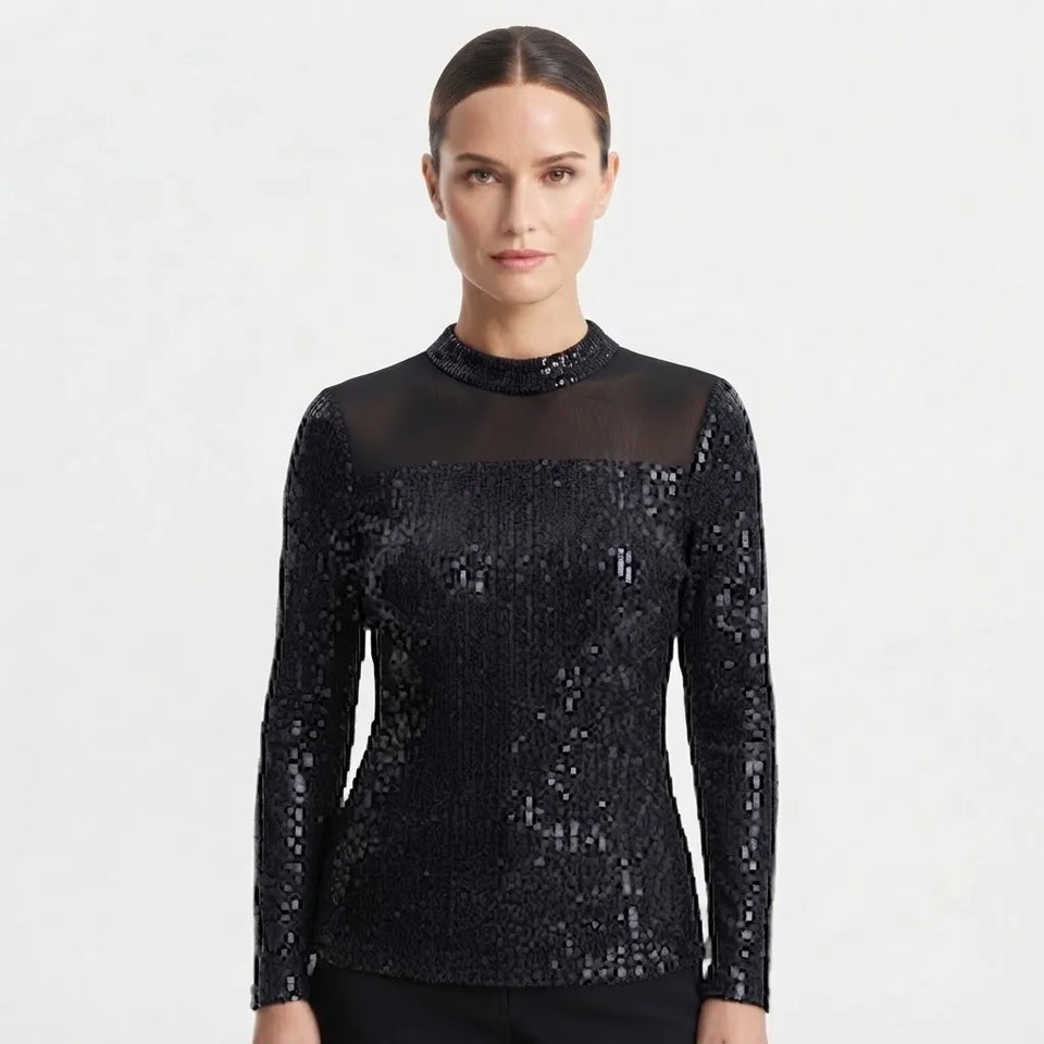 A woman modeling a black long-sleeve top with a sheer mesh mock neck and dense sequin embellishment across the bodice and sleeves, against a plain light gray studio background.