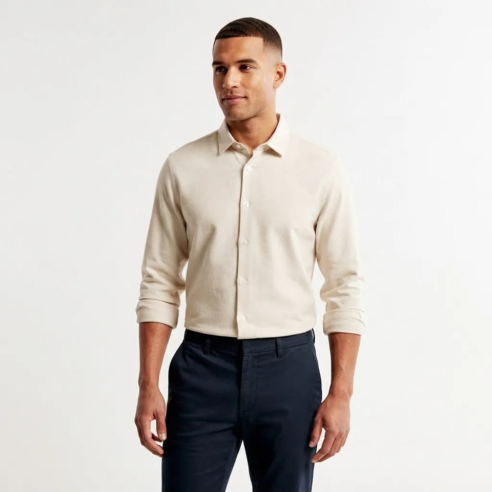 A man wearing a light beige long-sleeve knit button-up shirt and dark navy trousers, standing against a plain white studio background.