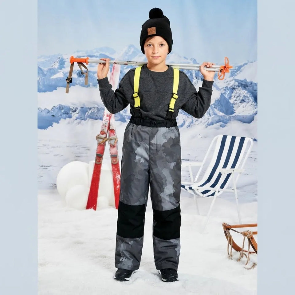 A child wearing a black beanie, dark gray long-sleeve top, and camouflage ski pants with black knee patches and yellow suspenders, holding ski poles in a snowy outdoor-style scene with skis and a chair.