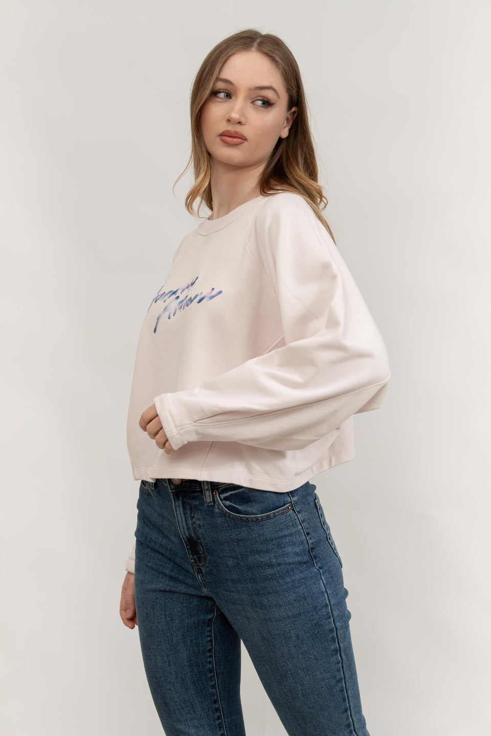 Woman wearing a beige sweatshirt with text and blue jeans on a plain background