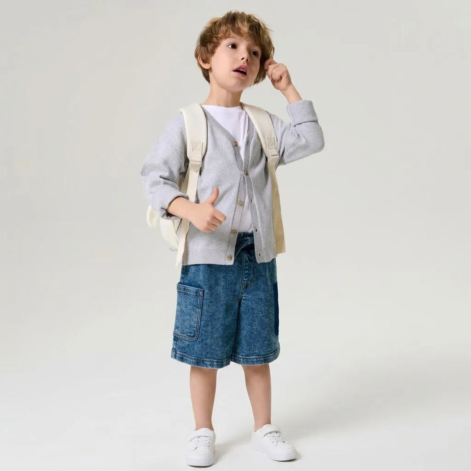 A young child stands against a light gray studio background wearing a light gray cardigan over a white shirt, blue denim shorts, white sneakers, and a cream backpack.