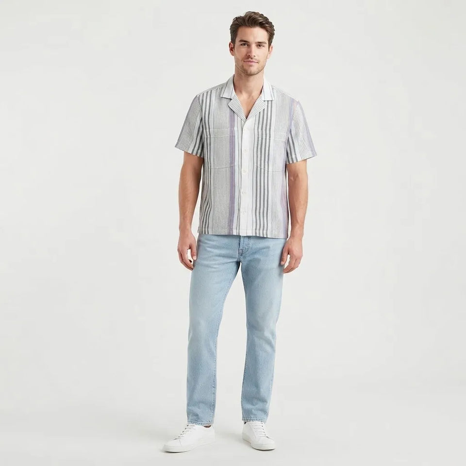 A man modeling a short-sleeve button-up shirt with vertical gray and white stripes, paired with light blue jeans and white sneakers, against a plain light gray studio background.