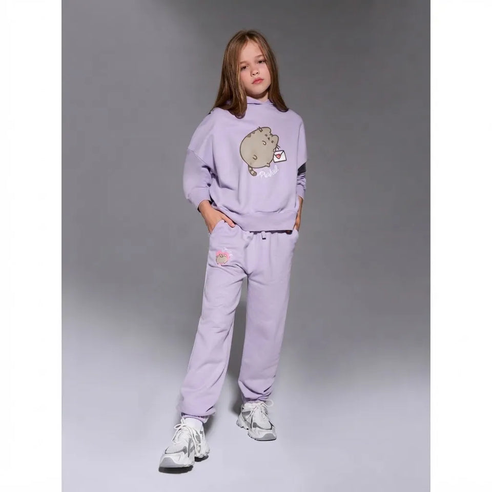 A young girl modeling a light purple/lavender Pusheen sweatshirt and matching sweatpants set, standing against a neutral gray studio background.