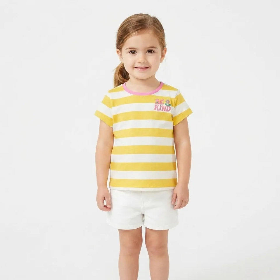 A young girl with light brown hair in a ponytail stands against a plain white studio background, wearing a yellow and white horizontally striped short-sleeve t-shirt with a pink collar and a small colorful graphic on the chest, paired with white shorts.