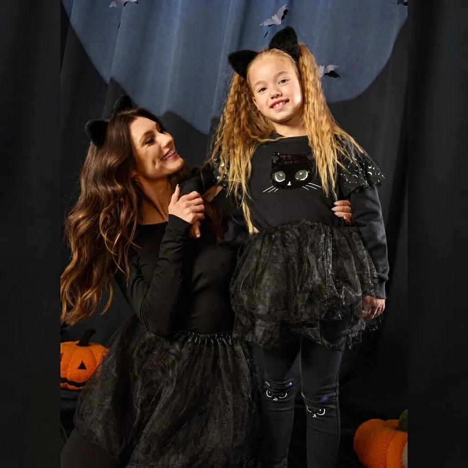 A woman and a young girl wearing matching black cat-themed Halloween costumes, standing in front of a dark curtain backdrop with pumpkins at the bottom.