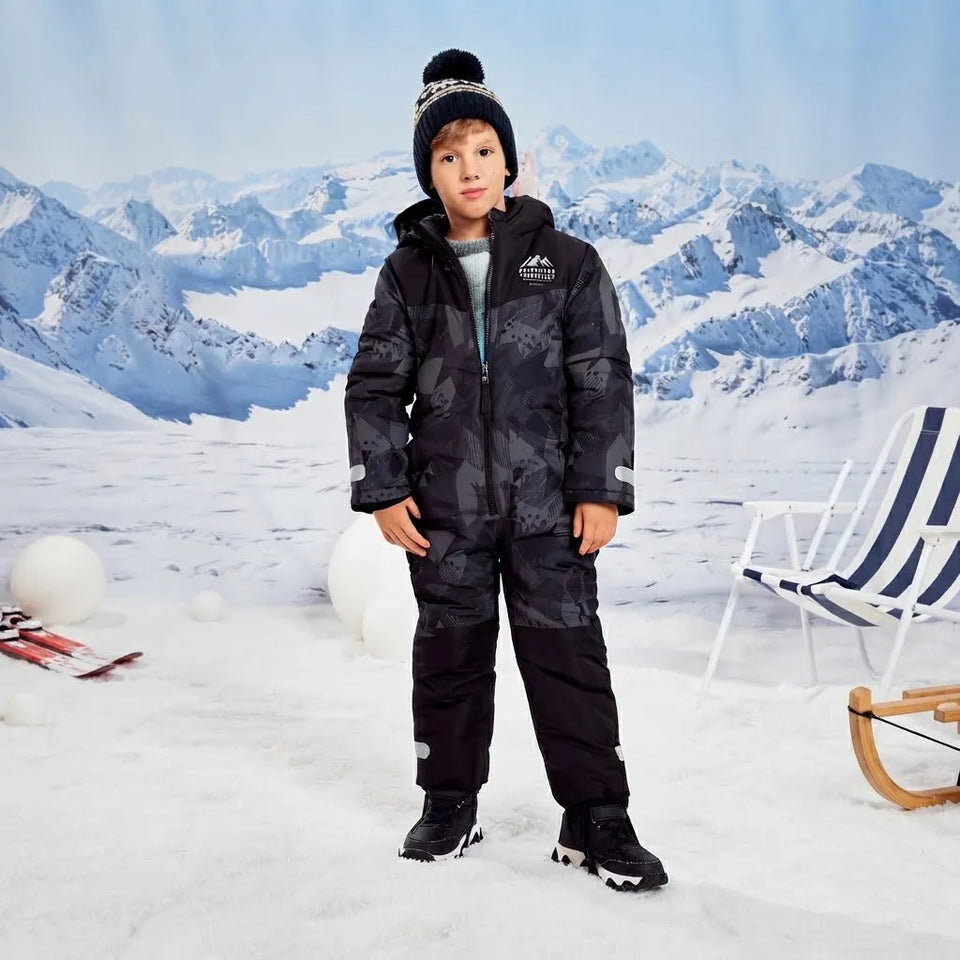 A young boy wearing a black and gray camouflage winter ski suit, black beanie with a pom-pom, and black ski boots, standing in a snowy landscape with a blue and white mountain backdrop. A blue and white striped beach chair is visible on the right.