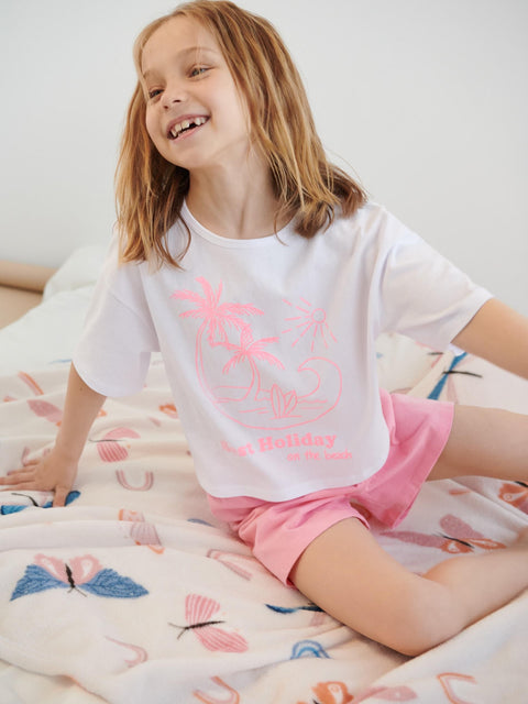 Young girl wearing a white t-shirt with pink graphics and pink shorts, sitting on a bed with a colorful blanket.