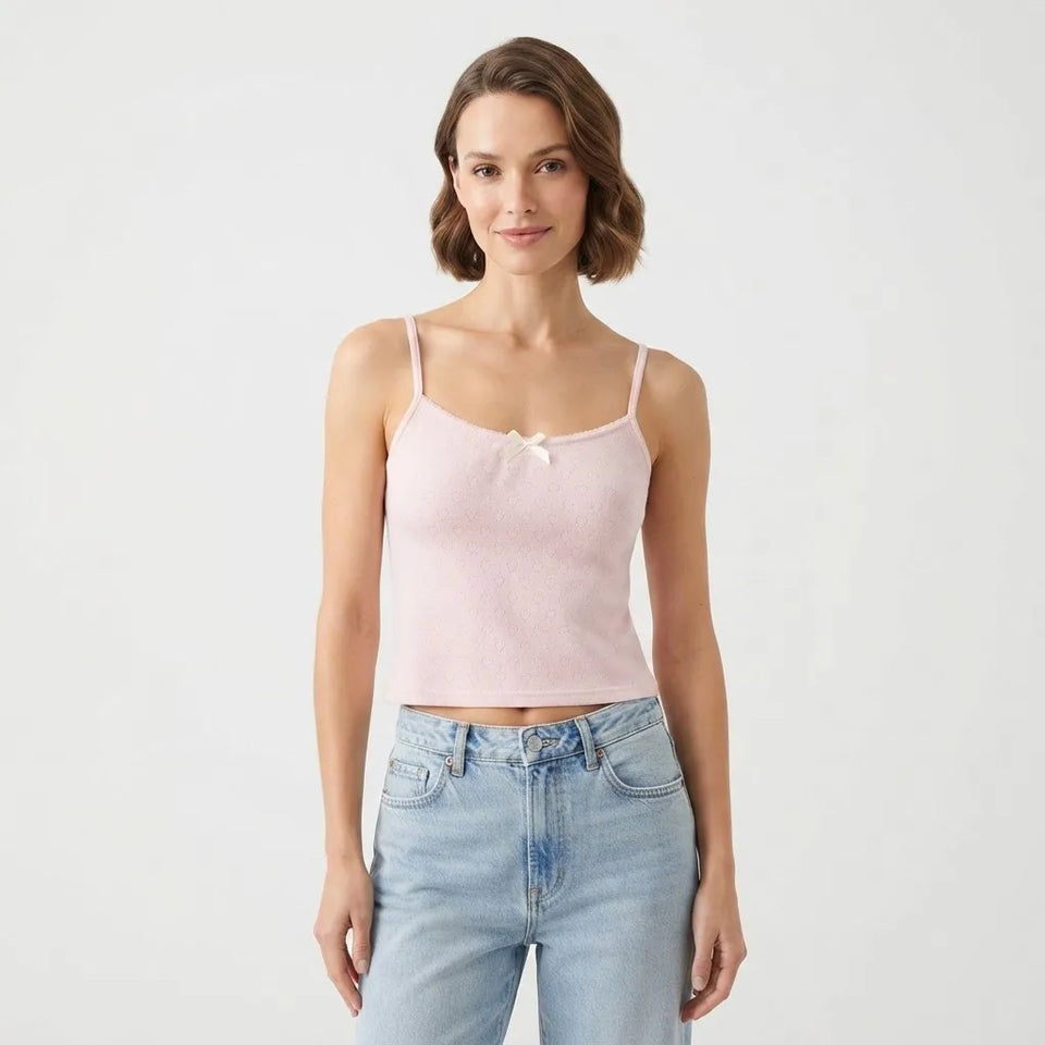 A woman modeling a light pink ribbed knit camisole top with thin straps and a small bow at the center chest, paired with light-wash blue jeans, against a plain light gray studio background.