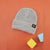 A light gray ribbed knit beanie with a folded cuff, featuring a small black rectangular patch with yellow text that reads 'LOOK UP'. The beanie is photographed on a solid orange background with decorative yellow and blue blocks in the lower right corner.
