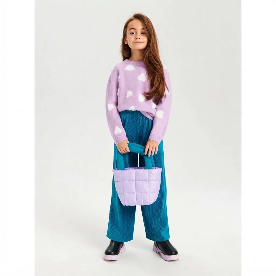 A young girl stands against a light gray studio background wearing a lavender fuzzy sweater with white cloud patterns, teal wide-leg pants, black boots, and holding a lavender quilted puffer handbag.