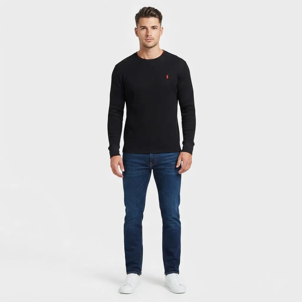 A man modeling a black long-sleeve crewneck sweater with a small red polo player logo on the left chest, paired with blue jeans and white sneakers, against a light gray studio background.
