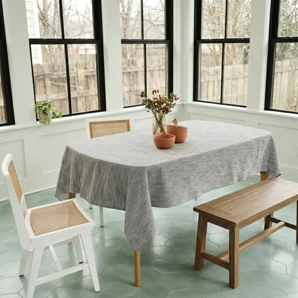 A gray textured tablecloth draped over a rectangular dining table in a bright room with large black-framed windows, a wooden bench, woven chairs, and small terracotta pots on the table.