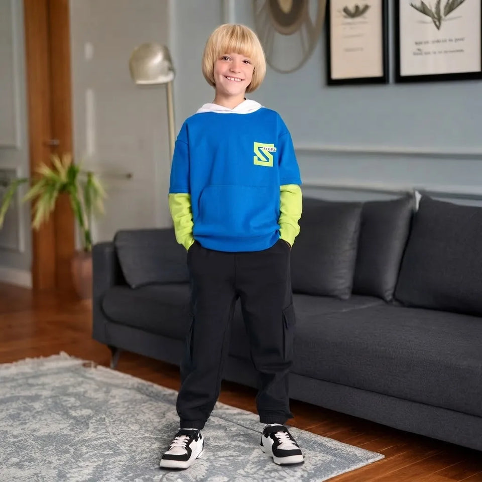 A young boy stands in a living room wearing a bright blue sweatshirt with neon green sleeves, black cargo pants, and white sneakers. The sweatshirt features a white hood and a yellow graphic logo on the left chest.