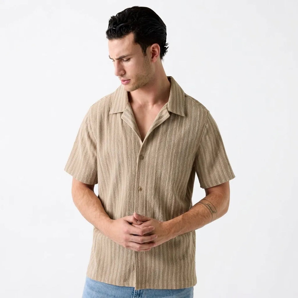 A man wearing a beige short-sleeve button-up shirt with vertical brown stripes, paired with blue jeans, photographed against a plain white studio background.