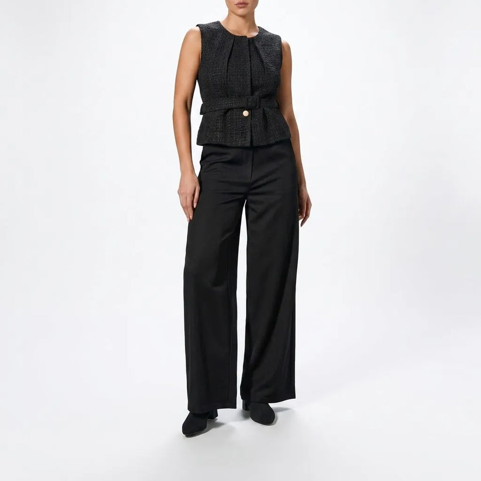 A model wearing a black sleeveless textured vest with a peplum waist and a single gold button, paired with black wide-leg trousers and black shoes, against a plain white studio background.
