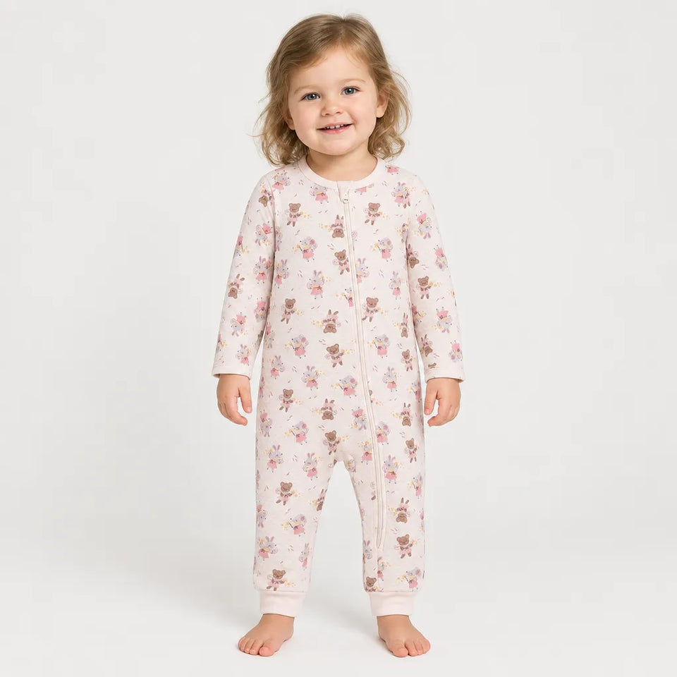 A smiling toddler stands barefoot against a plain light gray studio background, wearing a long-sleeve footless one-piece pajama or romper with a front zipper and an allover small teddy bear and floral print.