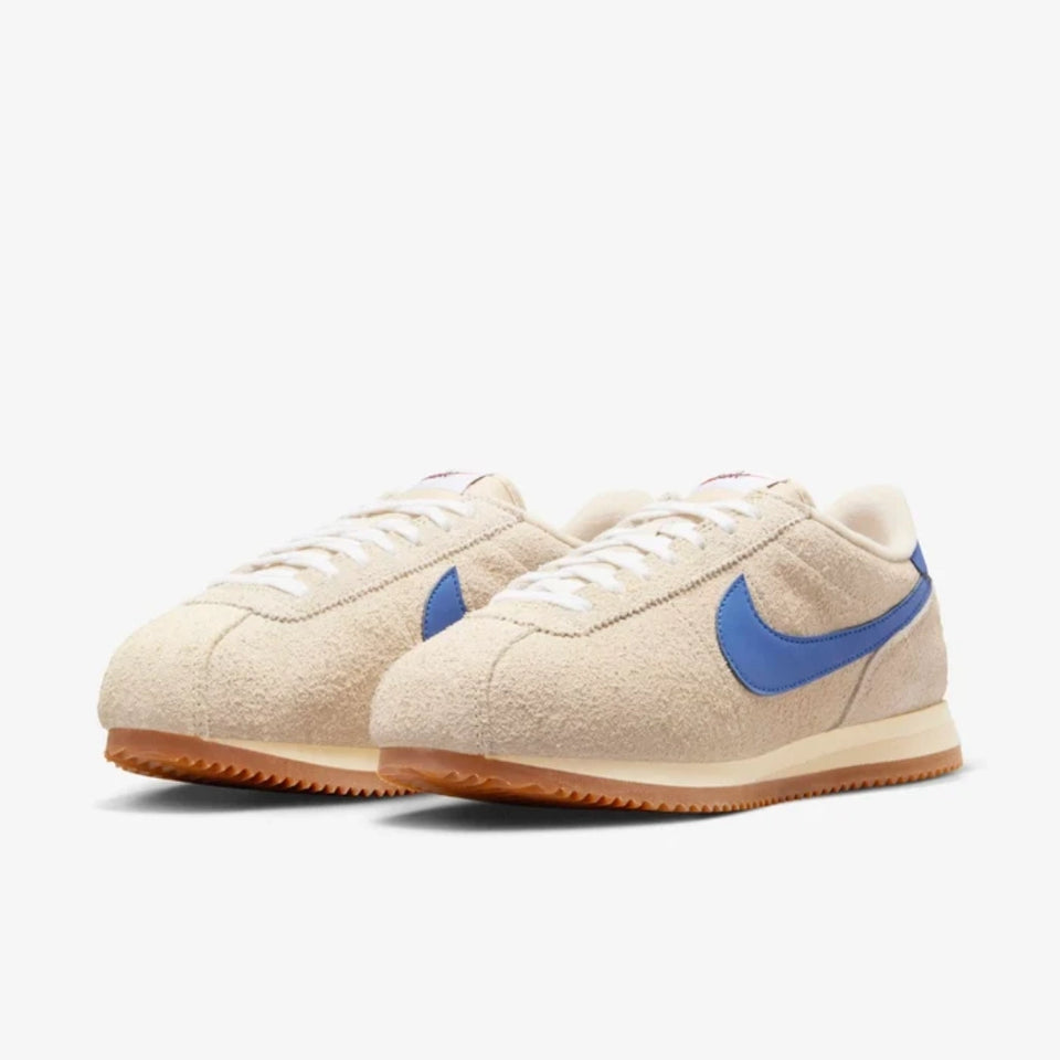 A pair of Nike Classic Cortez sneakers in beige suede with a vibrant blue Swoosh and a gum rubber sole, set against a clean white background.