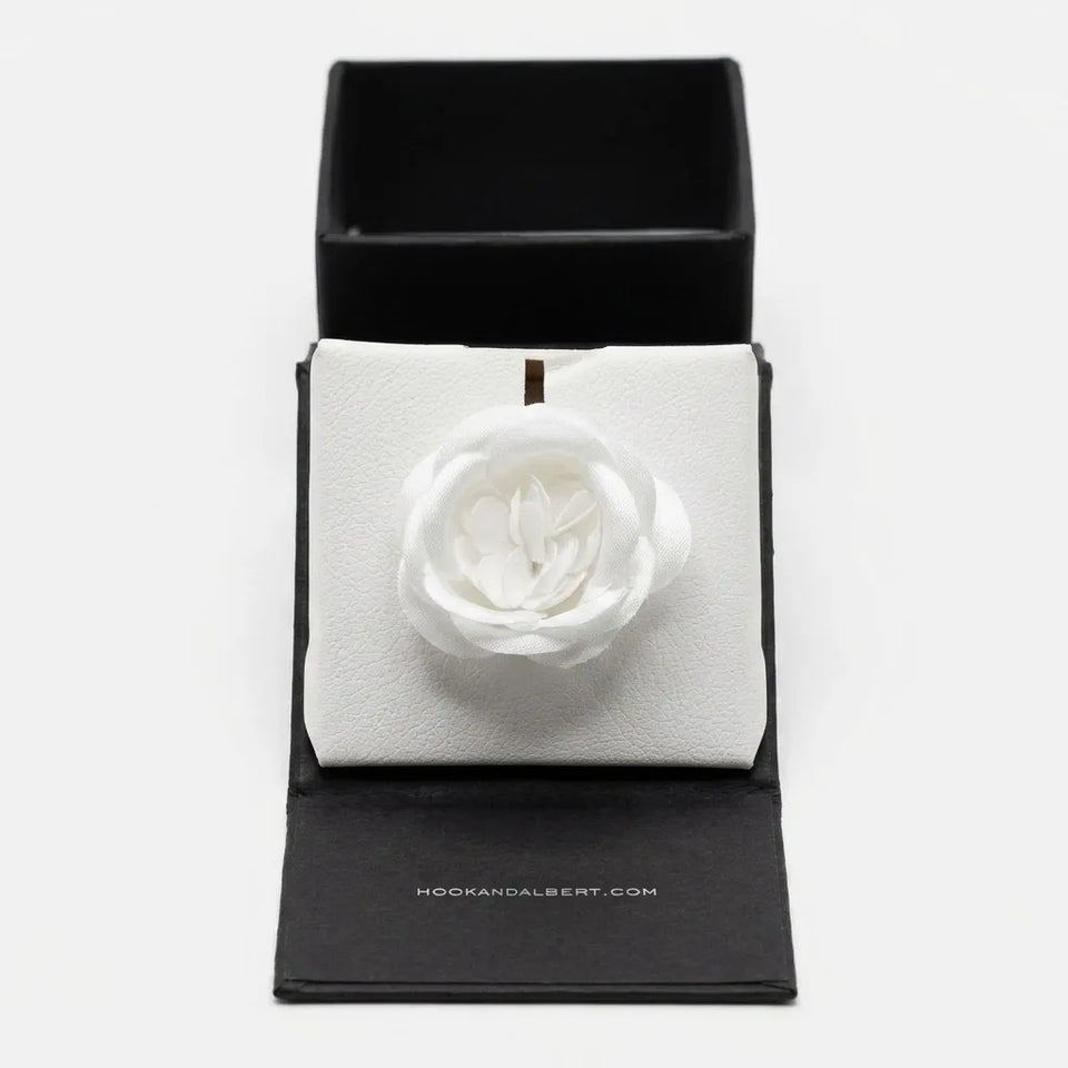 A white fabric flower brooch displayed on a white textured insert inside an open black presentation box. The brand name Hookandalbert.