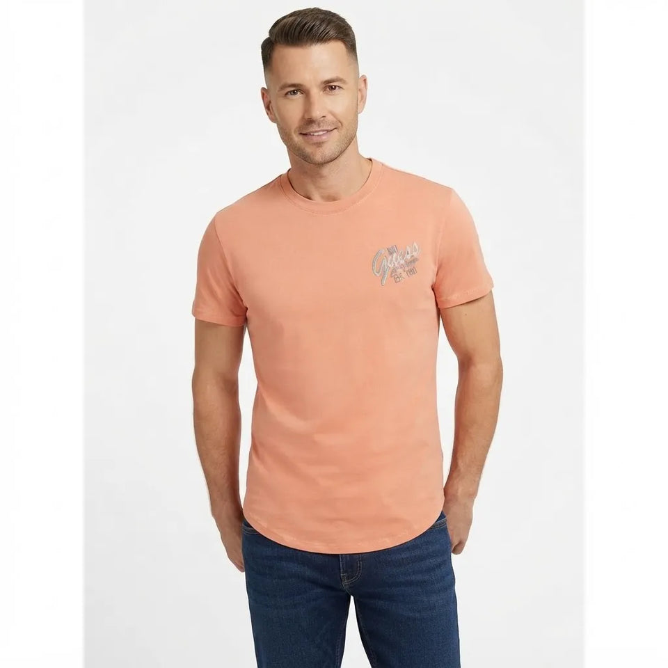 A man wearing a peach-colored short-sleeve t-shirt with a small graphic on the left chest, paired with dark blue jeans, standing against a light gray background.