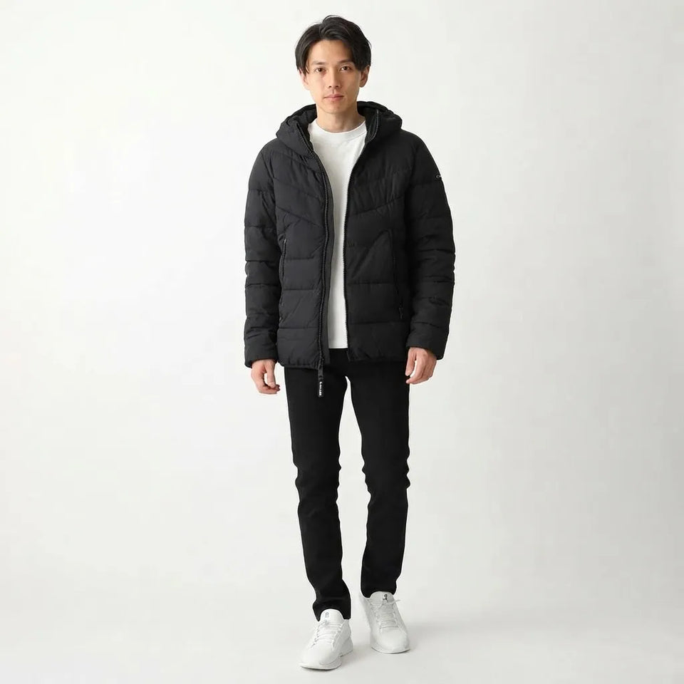 A man wearing a black hooded puffer jacket over a white t-shirt, black pants, and white sneakers, standing against a plain white studio background.