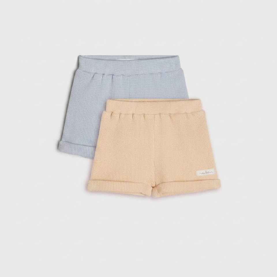 Two pairs of ribbed knit baby shorts, one in light blue and one in pale peach, displayed on a light gray background.
