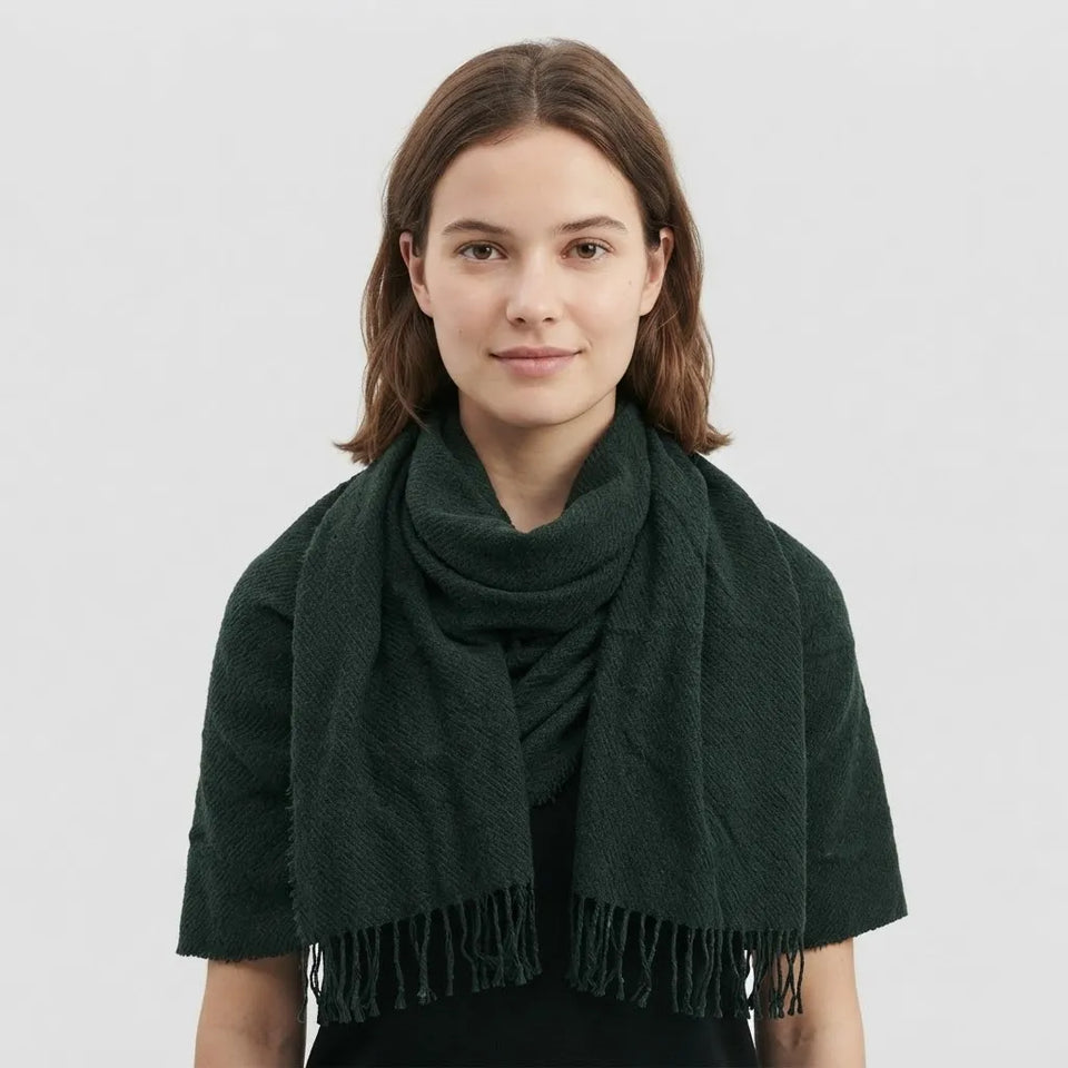 A woman with shoulder-length brown hair wearing a dark forest green textured knit scarf with fringe, styled over a black top against a light gray studio background.