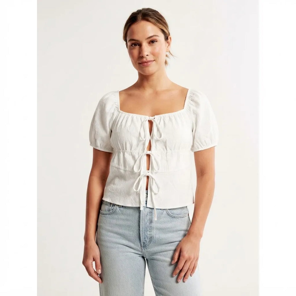 A woman modeling a white puff-sleeve top with a square neckline, gathered front, and tie closures, paired with light-wash jeans against a plain light background.
