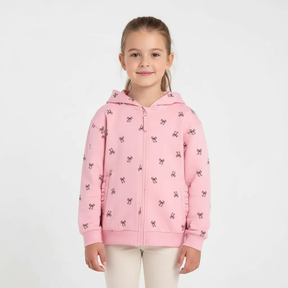 A young girl with her hair pulled back stands against a light gray studio background wearing a pink zip-up hoodie with a small black bow pattern and white pants.