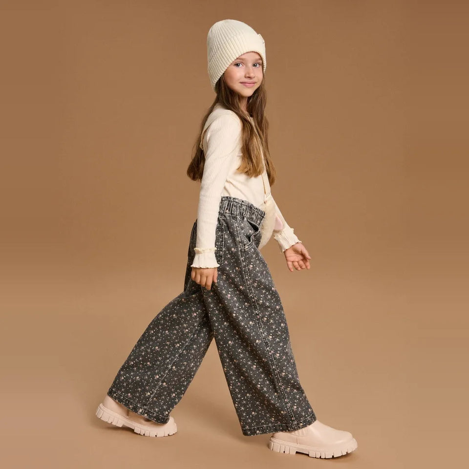 A young girl wearing a cream knit beanie, cream long-sleeve top, dark gray wide-leg pants with a small white floral pattern, and cream chunky platform shoes, walking against a solid tan studio background.