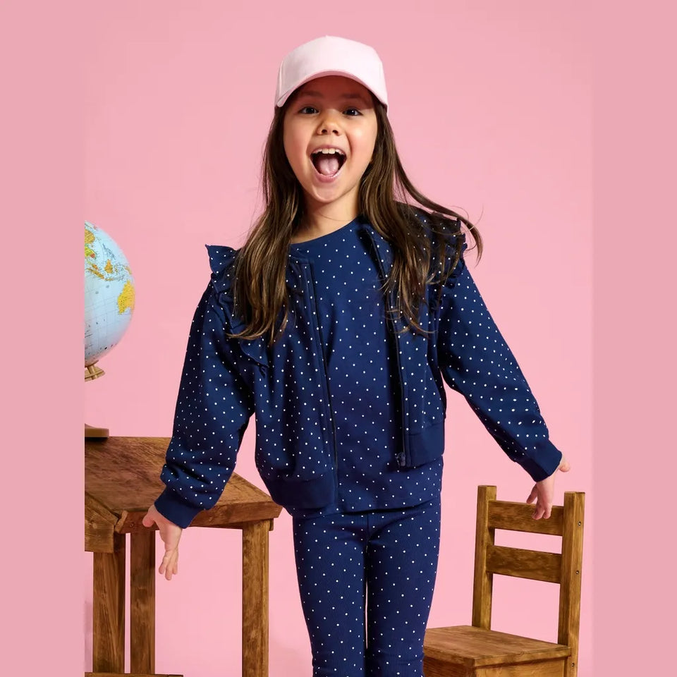 A young girl with long brown hair wearing a navy blue and white polka dot matching set, consisting of a long-sleeve top with ruffle details and matching pants, posing in front of a pink background with wooden chairs and a globe.