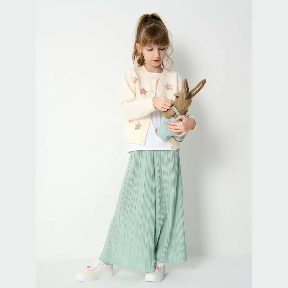 A young girl stands against a plain white studio background wearing a cream cardigan with star patches, a white top, and light green pleated wide-leg pants, while holding a stuffed bunny.