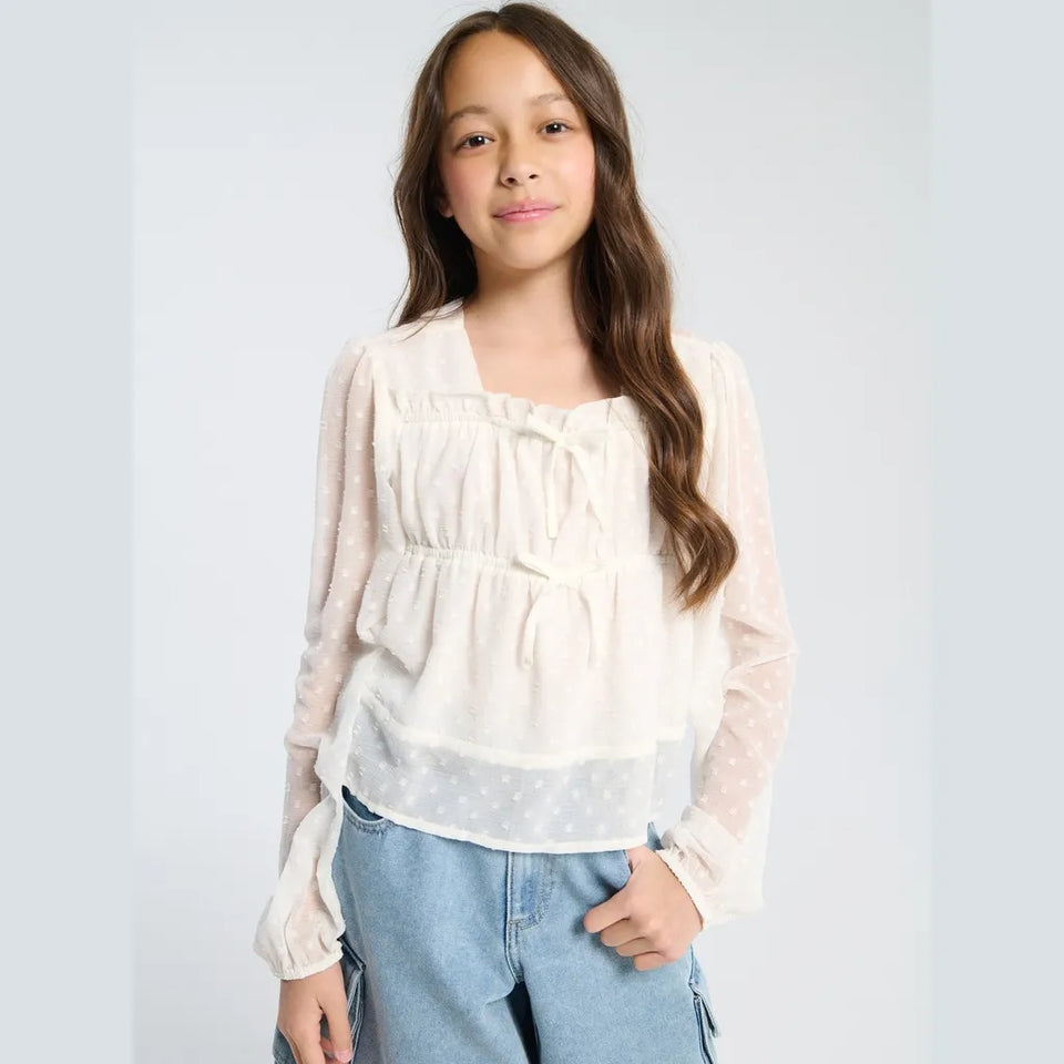 A young girl with long brown hair wearing a cream-colored long-sleeve blouse with sheer polka dot sleeves and a ruffled tiered bodice, paired with light blue jeans, against a light gray studio background.