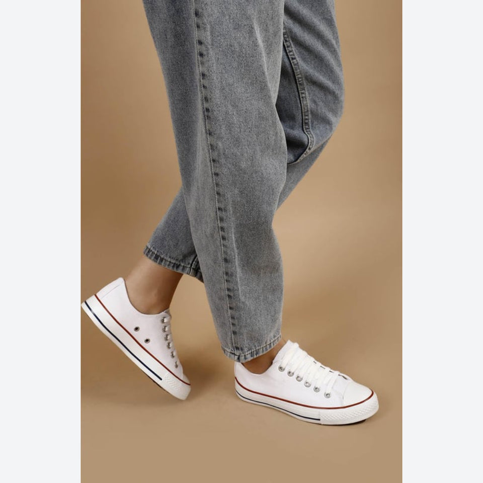 White low-top canvas sneakers with silver eyelets and a thin red stripe, shown on a model wearing gray jeans against a solid tan background.