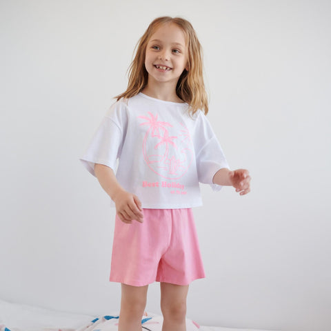 Young girl wearing a white t-shirt with pink text and pink shorts on a plain background