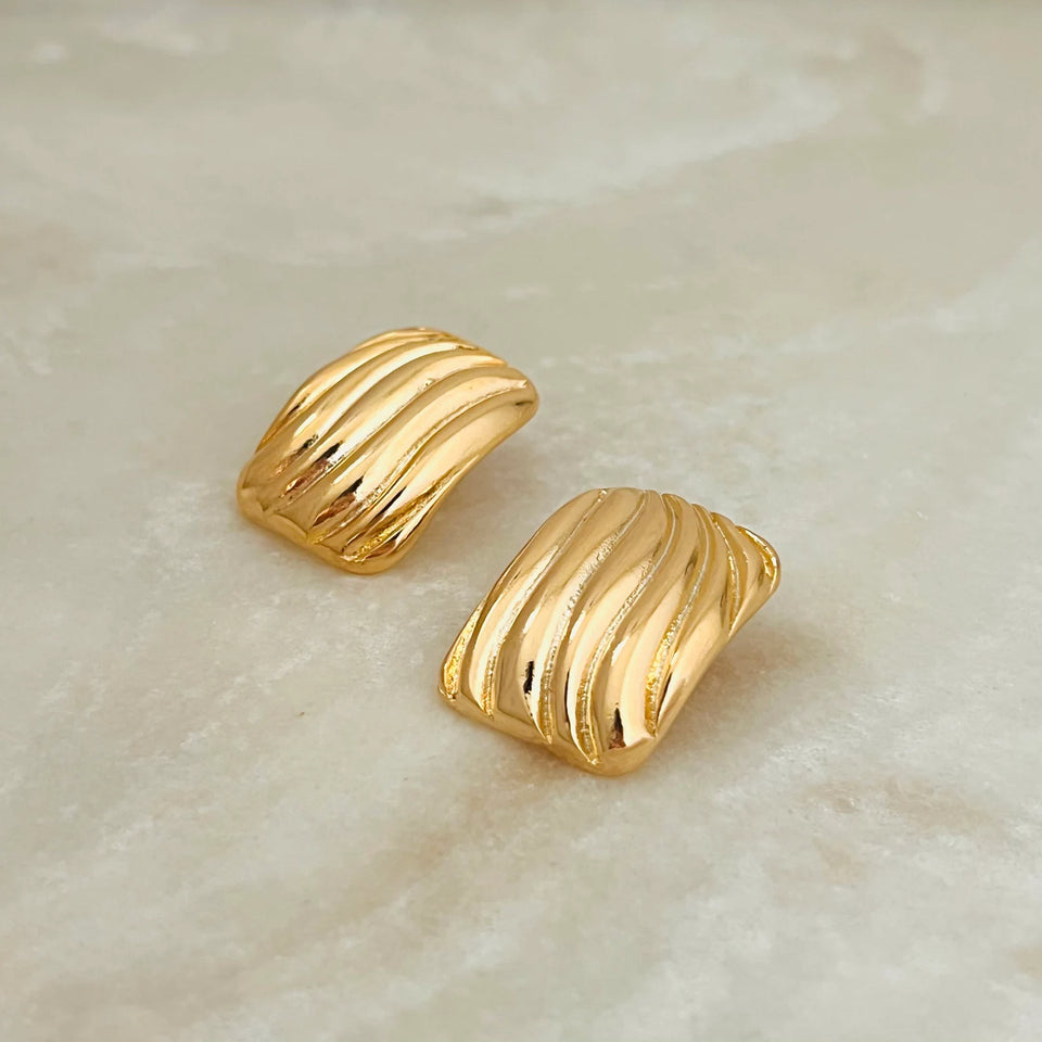 A pair of gold-toned textured stud earrings with a ribbed, wave-like pattern, displayed on a light beige marble surface.