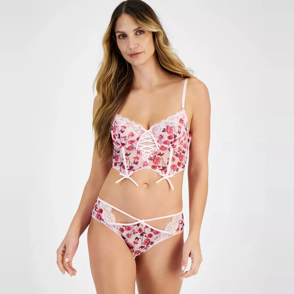 A woman modeling a two-piece lingerie set featuring a pink and red floral lace bralette with white straps and matching lace panties, set against a plain light gray studio background.