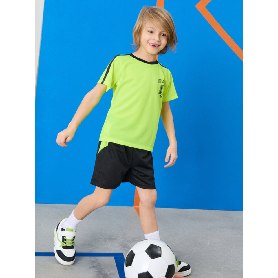 A young boy wearing a neon green soccer t-shirt with black trim, black shorts, and white sneakers with green accents, standing next to a soccer ball in front of a blue wall.