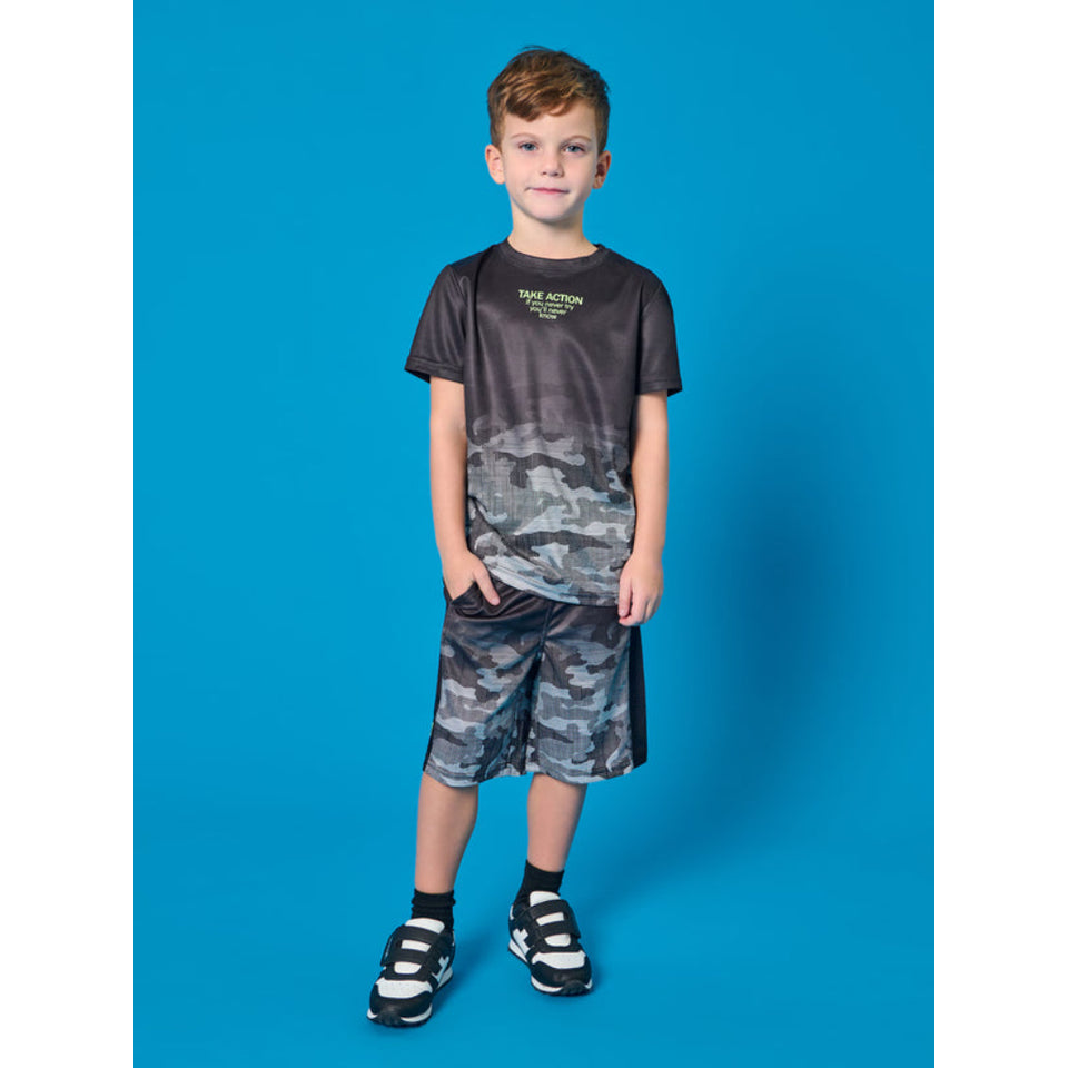 A young boy modeling a matching two-piece athletic set consisting of a dark gray gradient and camouflage print t-shirt and shorts, paired with black and white sneakers, against a solid blue studio background.