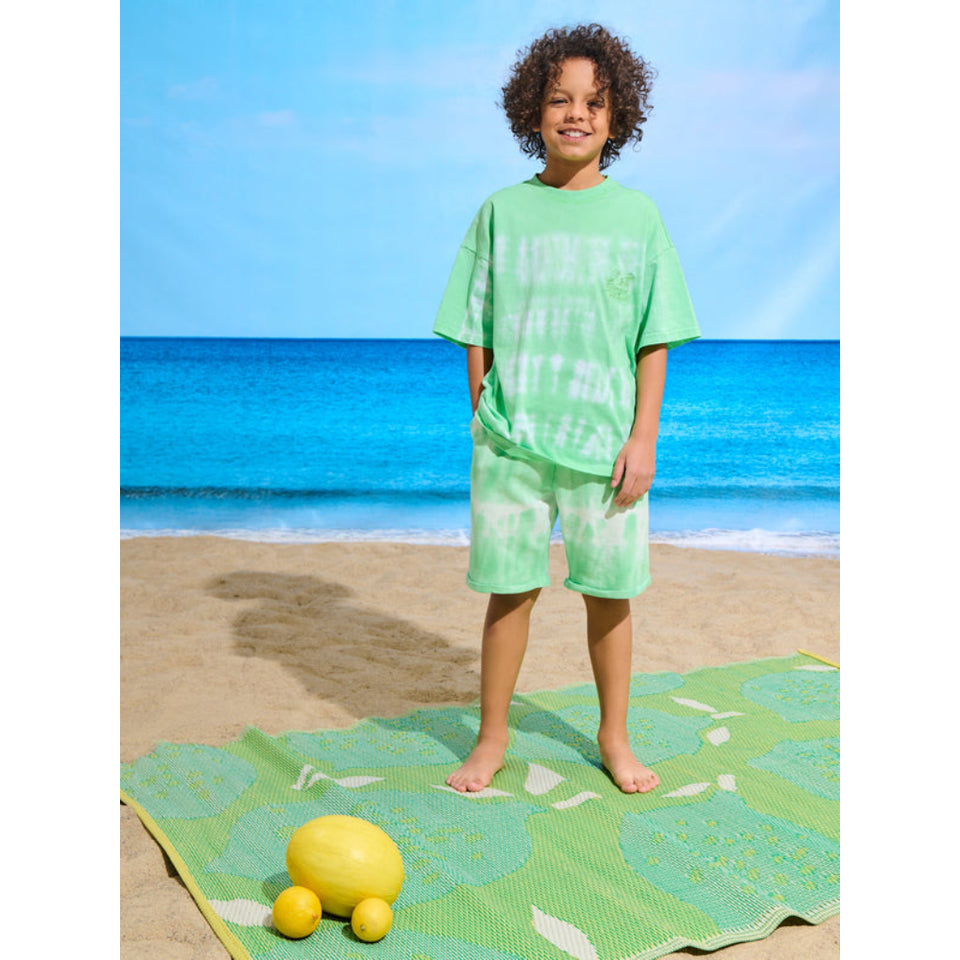A child stands on a beach towel at the beach wearing a matching light green t-shirt and shorts set. The towel has a green and yellow tropical leaf pattern, and there are lemons on the sand in the foreground.