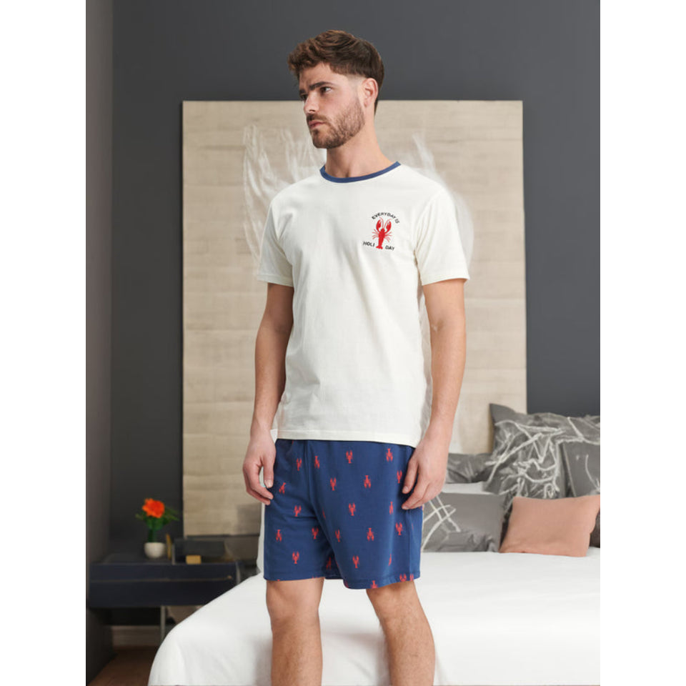 A man wearing a two-piece pajama set consisting of an off-white short-sleeve t-shirt with a red lobster graphic and navy blue shorts with a red lobster pattern, standing in a bedroom setting.