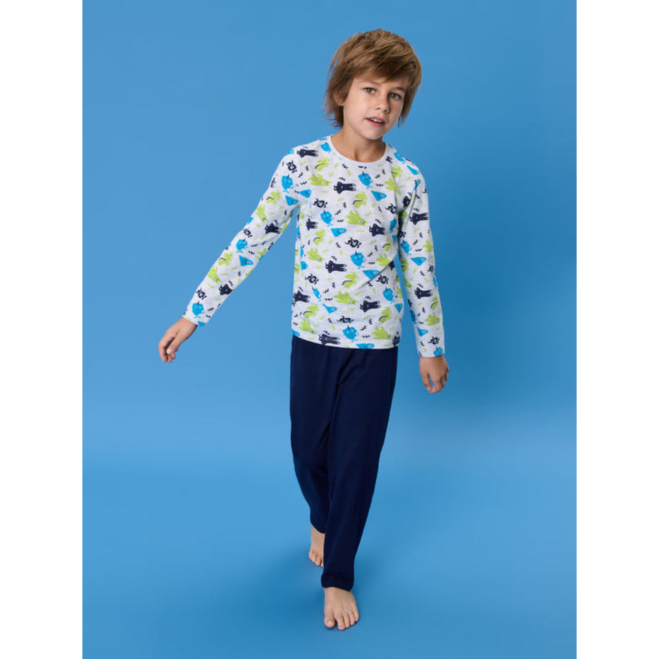 A young boy modeling a long-sleeve pajama top with a colorful dinosaur print on a white background, paired with dark navy pants, against a solid blue studio backdrop.