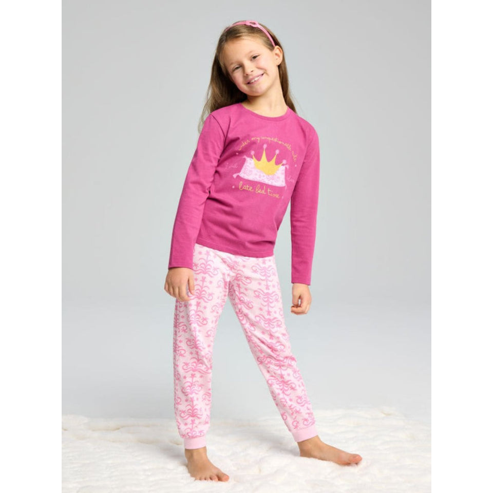 A young girl stands barefoot on a white textured rug against a light gray studio background, wearing a pink long-sleeve pajama top with a crown graphic and matching pink patterned pajama pants.