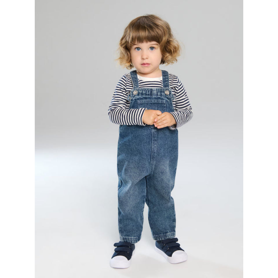A toddler standing against a light gray studio background, wearing blue denim overall with a black-and-white striped long-sleeve shirt underneath and dark shoes with white soles.
