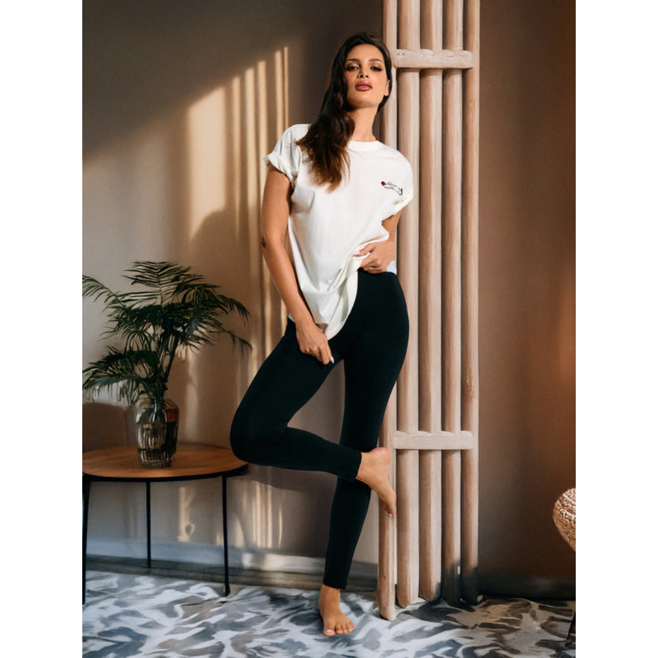 A woman posing in a white short-sleeve t-shirt with a small graphic on the chest and black leggings, standing in a styled room with warm lighting and a wooden slatted screen.