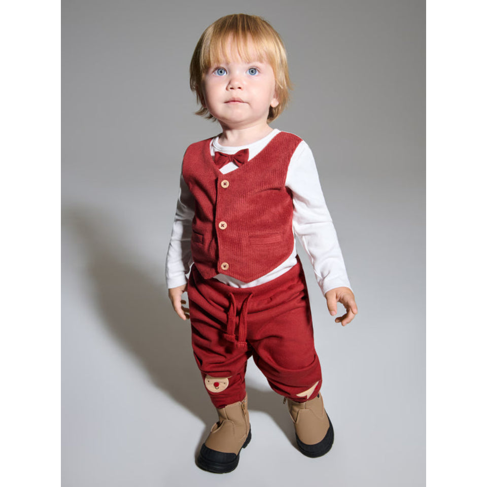 A toddler standing against a plain gray studio background, wearing a red corduroy vest and matching corduroy pants over a white long-sleeve shirt, with a small red bow tie and tan boots.